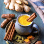 Cup of golden turmeric latte with cinnamon stick, fresh turmeric root, ginger, and cardamom on rustic wooden table.