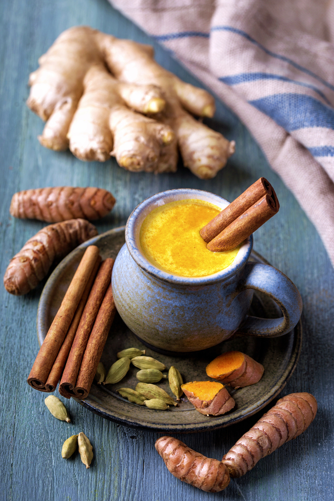 Cup of golden turmeric latte with cinnamon stick, fresh turmeric root, ginger, and cardamom on rustic wooden table.