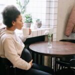 Pregnant woman sitting in a cozy café, gently cradling her belly with one hand while sipping from a white mug with the other, surrounded by natural light and potted plants — capturing a calm moment during her transition away from coffee.