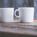 Two plain white ceramic coffee mugs sit side by side on a rustic wooden surface, with a soft-focus, neutral-toned background — symbolizing a side-by-side comparison between two coffee alternatives.