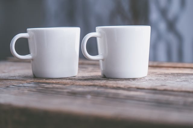 Two plain white ceramic coffee mugs sit side by side on a rustic wooden surface, with a soft-focus, neutral-toned background — symbolizing a side-by-side comparison between two coffee alternatives.